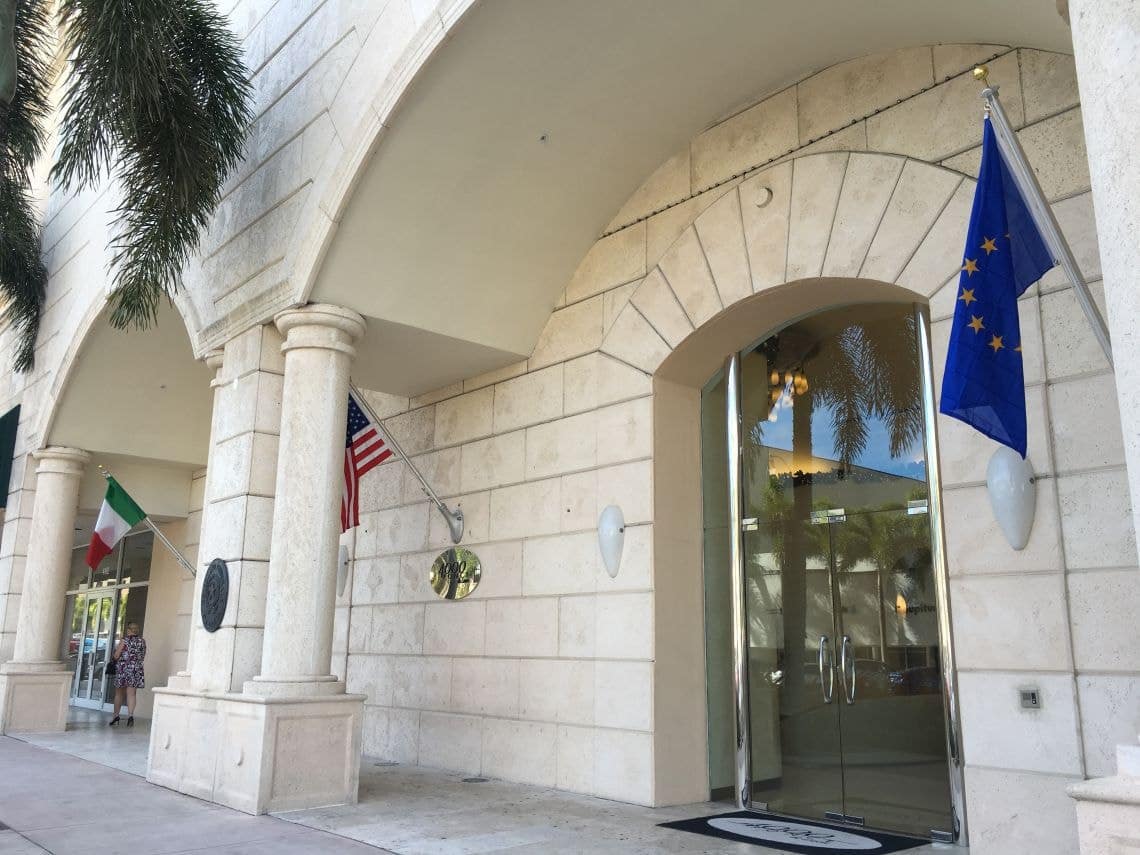 The image depicts the exterior of the Italian Consulate General located at 4000 Ponce de Leon in Coral Gables, Florida, featuring both the Italian and American flags prominently displayed. The professional architecture reflects the consulate's role in international cooperation and foreign affairs.